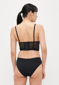 Schwarzer BH mit transparenter Rückenpartie, verstellbaren Trägern und Hakenverschluss. Kombiniert mit passenden, hoch taillierten schwarzen Slips mit Spitzenbesatz.