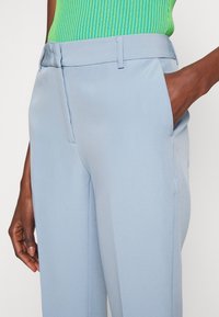 Gros plan d'un pantalon bleu clair ajusté avec poches latérales, porté avec un haut vert côtelé ; une main repose dans la poche, l'autre le long du corps.