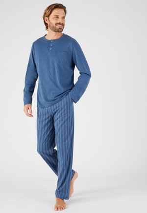 Uomo in piedi a piedi nudi che indossa una maglia henley blu a maniche lunghe e pantaloni del pigiama a righe blu su uno sfondo semplice.