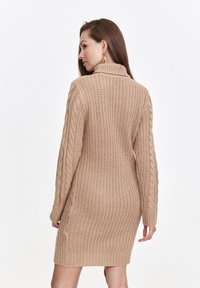 Strickkleid in Beige mit gerippter Textur und Kabelstrickärmeln. Besitzt einen hohen Kragen und eine figurbetonte Silhouette.