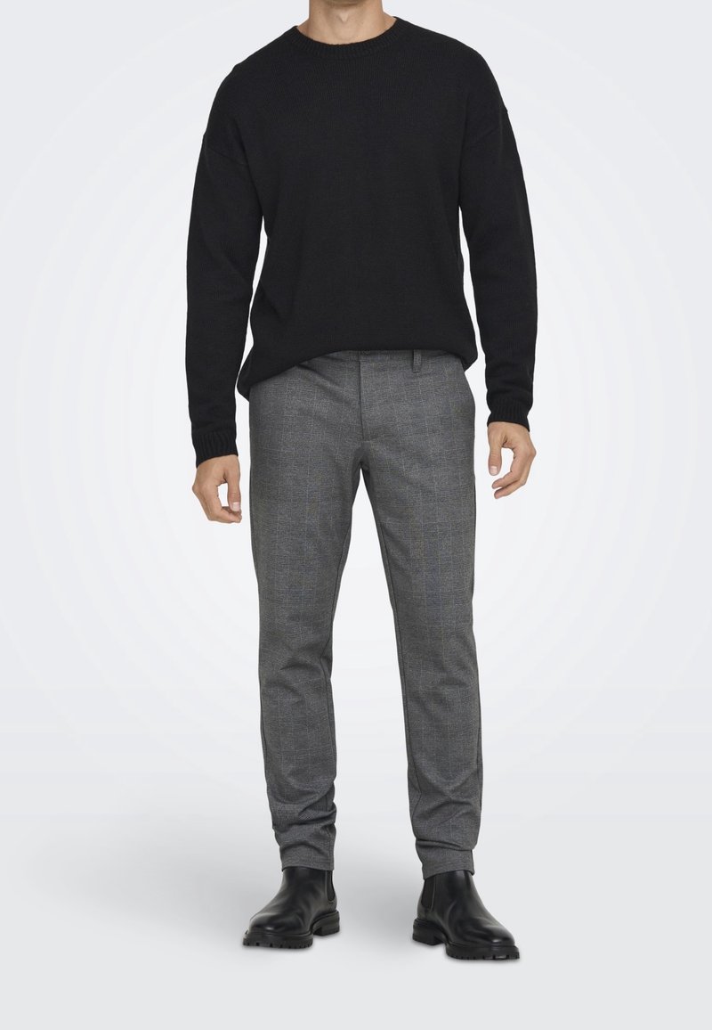 Homme portant un pull noir col rond, un pantalon slim gris à carreaux et des bottines Chelsea en cuir noir, debout devant un fond blanc.