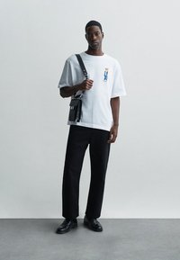 Bărbat îmbrăcat cu o cămașă albă cu grafică, pantaloni negri, pantofi din piele negri și cărând o geantă de umăr neagră, stând în fața unui perete simplu de culoare deschisă.