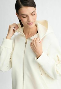 Witte zip-up hoodie met een capuchon, grote zakken, gouden ritsdetails en geribde manchetten. Gladde textuur met een relaxte pasvorm.