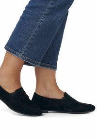 Scarpe slip-on in suede nero con tacco basso, caratterizzate da una fascia decorativa sul davanti e una texture liscia e opaca. Indossate con jeans cropped.