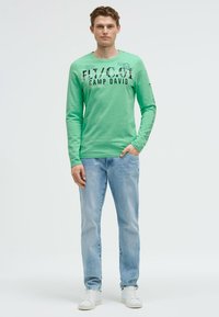Grüner Langarm-T-Shirt mit Grafikdruck und Text; kombiniert mit hellblauen Jeans und weißen Sneakers; lässiger Schnitt und leichter Stoff.