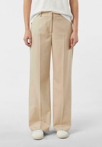 Beżowe spodnie o szerokich nogawkach wykonane z teksturowanej tkaniny, z płaskim przodem, szlufkami na pasek i dopasowanym krojem. Noszone z białymi trampkami.