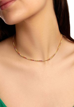 Gros plan sur le cou d'une femme portant un collier fin de pierres précieuses colorées sur une peau nue et un vêtement vert.