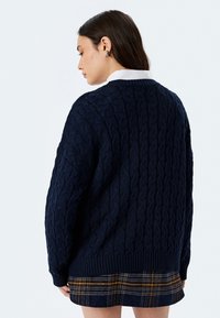 Maglione blu navy a maglia cable, con orlo e polsini a costine, caratterizzato da motivi texturizzati. Indossato sopra una gonna a quadri con tonalità gialle e blu navy.