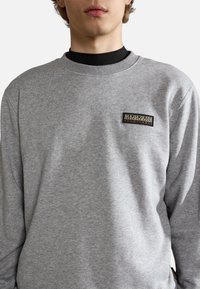 Sweat-shirt gris à manches longues, col rond, avec un patch logo texturé sur la poitrine. Le tissu semble doux avec une coupe décontractée.