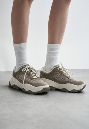 Osoba nosząca białe skarpety za kostkę oraz masywne beżowo-kremowe sneakersy na szarej podłodze na tle jasnego tła.