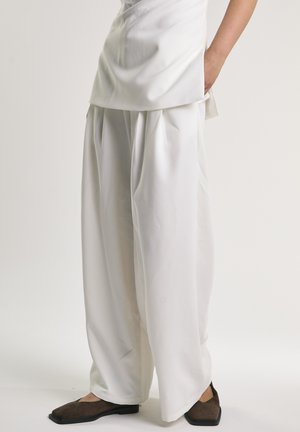 Löysät valkoiset housut, joissa on laskokset, yhdistetty valkoiseen yläosaan ja tummanruskeisiin neliökärkiin mataliin kenkiin.