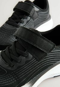 Chaussures de sport noires avec une tige en mesh, des rayures texturées, une sangle Velcro et des lacets. La semelle en caoutchouc blanche offre un contraste.