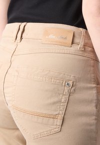 Beige jeans med en glatt tekstur, med et lærmerke, baklommer med sydd detaljer og en tettsittende design.