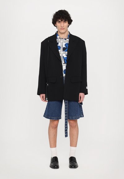 Black oversized wool blazer, bold printed blue and white shirt, denim shorts with raw hems, and black shoes with socks. Design is contemporary.