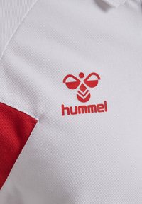 Vit pikétröja med en röd chevrondesign och en röd "hummel"-logotyp. Tyget har en texturerad yta.