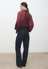 Bluză transparentă de burgundy cu mâneci bufante, asortată cu blugi evazați din denim închise, cu talie înaltă, având cusături subtile și buzunare la spate.
