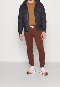 Svart quiltad pufferjacka med huva, brun tröja, bruna sweatpants och vita sneakers med orangea accenter. Har dragsko och ribbade mudd.