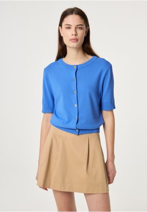 Vrouw die een felblauwe, korte mouwen, met knopen, vest draagt en een beige geplooide rok, staande tegen een effen witte achtergrond.