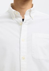 Gros plan d'une personne portant une chemise blanche boutonnée avec un col boutonné et une poche poitrine, montrant une partie du cou et du haut de la poitrine.