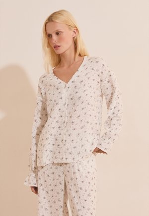 Donna bionda con top bianco a maniche lunghe abbottonato e pantaloni larghi abbinati con piccola stampa floreale, in piedi su uno sfondo chiaro uniforme.