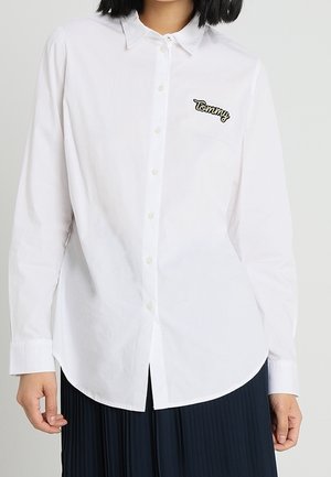 Personne portant une chemise blanche à boutons avec "Tommy" brodé sur la poitrine, associée à une jupe plissée sombre.