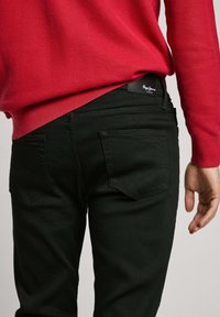Black jeans with back pockets and subtle stitching details, paired with a red textured knit sweater. The jeans have a fitted shape.