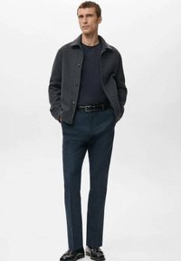 Mann trägt dunkelgraue Jacke, schwarzes Hemd, dunkle blaue Hose und schwarze Schuhe, steht mit den Händen in den Taschen vor einfachem Hintergrund.