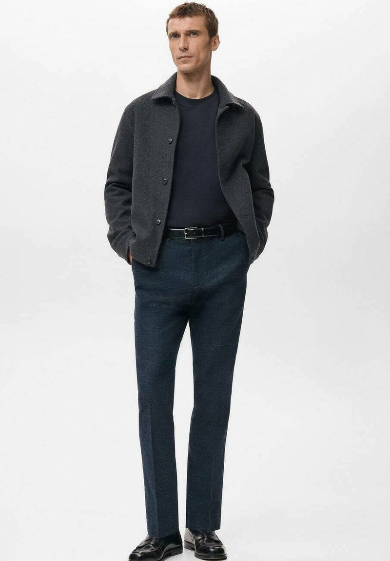 Homme portant une veste gris foncé, une chemise noire, un pantalon bleu foncé et des chaussures noires, debout les mains dans les poches devant un fond uni.