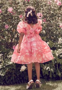 Roza cvetlična obleka z napihnjenimi rokavi, poudarjenim pasom in večplastno krilom. Značilnosti so velika pentlja na hrbtni strani in bele sandale s trakovi.