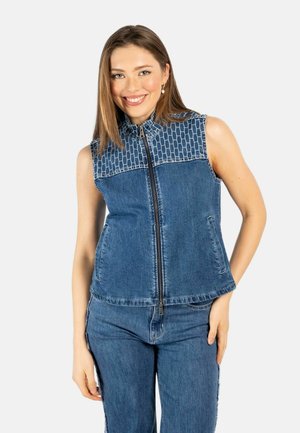 Smilende kvinne med lyst brunt hår iført ermeløs denimvest med glidelås og mønstret skulderparti, sammen med matchende denimjeans.