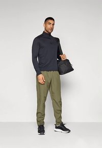 Moški, oblečen v črno majico z dolgimi rokavi, olivno zelene hlače, črne superge, z rjuho črne telovadne torbe preko enega ramena, stoji pred enobarvnim ozadjem.