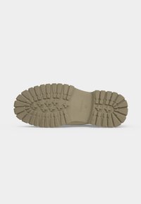 Semelle de botte en caoutchouc beige avec des crampons profonds et robustes et des motifs en forme de croix, vue de dessous sur fond blanc uni.