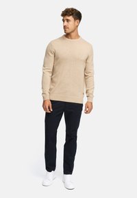 Suéter de punto beige con cuello redondo y puños acanalados, combinado con pantalones azul marino y zapatillas blancas, exhibido en un modelo masculino.