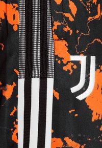 Černé tričko s oranžovým skvrnitým vzorem, obsahující texturovaný design, černou síťovinu a bílé pruhy, a bílou vyšitou nášivku.