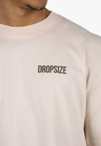 Világos rózsaszín pamut póló, kerek nyakkivágással. A bal oldalon sötétszürke "DROPSIZE" felirat olvasható. Sima tapintású és laza fazonú.