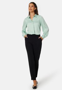 Světlezelená saténová blůza s knoflíkovým zapínáním a balónovými rukávy, v kombinaci s oblekovými černými kalhotami a černými plochými botami.