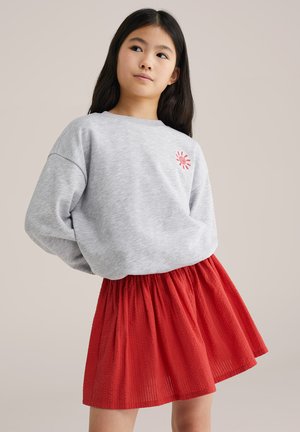 Jeune fille aux longs cheveux bruns portant un sweat-shirt gris clair avec un petit motif de soleil rouge et une jupe rouge texturée, debout devant un fond neutre.