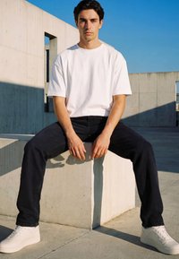 Young man wearing white t-shirt, black jeans, and white sneakers sitting on a concrete block against blue sky and modern concrete buildings.