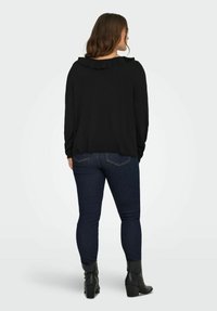 Žena okrenuta leđima, obučena u crnu bluzicu s nabranim ovratnikom, tamnoplave traperice i crne čizme s potpeticom na ankletima, na jednobojnoj svijetloj pozadini.