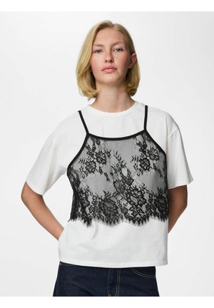 Blondínka v bielom tričku s čiernym kvetinovým čipkovaným tielkom cez neho, stojaca s jednou rukou na bokoch.