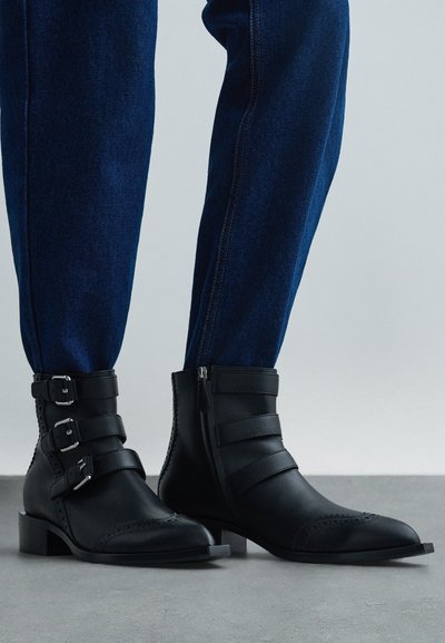 Kājas, kas tērptas tumši zilos džinsos, kombinējot tos ar melnām ādas potīšu zābakām, kurām ir trīs sprādzes un sānu rāvējslēdzis.