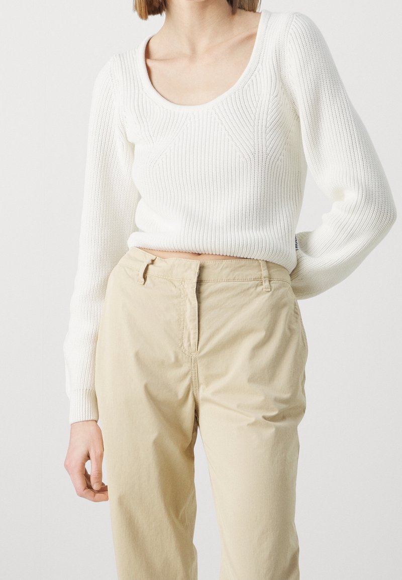 Femme portant un pull à manches longues blanc en laine tricotée et un pantalon taille haute beige sur un fond blanc uni.