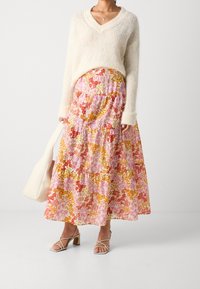 Femme portant un pull crème à col en V duveteux, une jupe maxi fleurie rose-orange, des sandales à talons nu et à lanières, tenant un sac à main blanc.