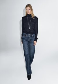 Black turtleneck sweater with long sleeves, paired with high-waisted blue denim jeans. Model wears black heeled boots and a silver pendant necklace.