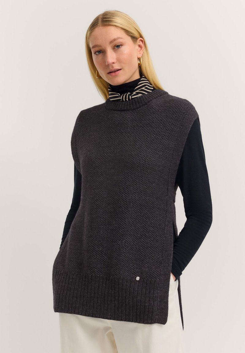 Chunky knit dark gray sleeveless sweater with high neckline, side slits, and ribbed hem, layered over a black long-sleeve shirt.