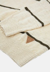 Pull en maille crème avec des rayures vert foncé. Présente un tissu texturé et une étiquette triangulaire en cuir marron avec "Guess" embossé.