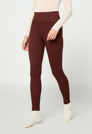 Leggings in Dunkelbraun, getragen von einer stehenden Person mit cremefarbenen Socken und einem langärmeligen beigen Oberteil, das oberhalb der Brust gekürzt ist.