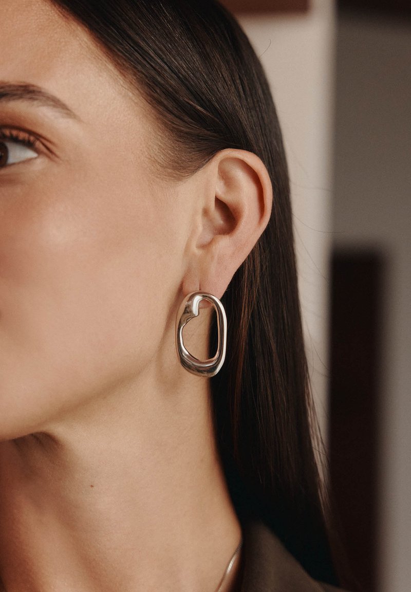 Orecchino a cerchio in argento con una forma aperta e astratta. Superficie liscia con una finitura riflettente. Posizionato sull'orecchio di un modello, che mette in risalto il suo design.