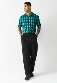 Cămașă cu mâneci scurte, în carouri verzi, cu nasturi, asortată cu pantaloni negri cu picioare largi. Modelul stă cu mâinile în buzunare, purtând pantofi negri.