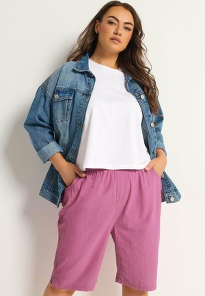 Vrouw met een spijkerjack, wit T-shirt en losse roze short, staand met handen in de zakken tegen een effen lichte achtergrond.